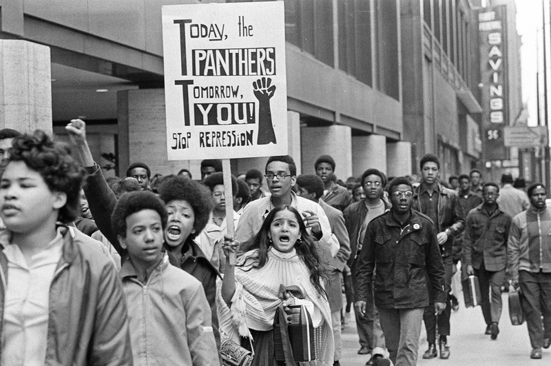 Black and white image of protesters marking down the street towards Daley Center. One protester is holding a sign that says "Today, the Panthers Tomorrow, You Stop Repression".