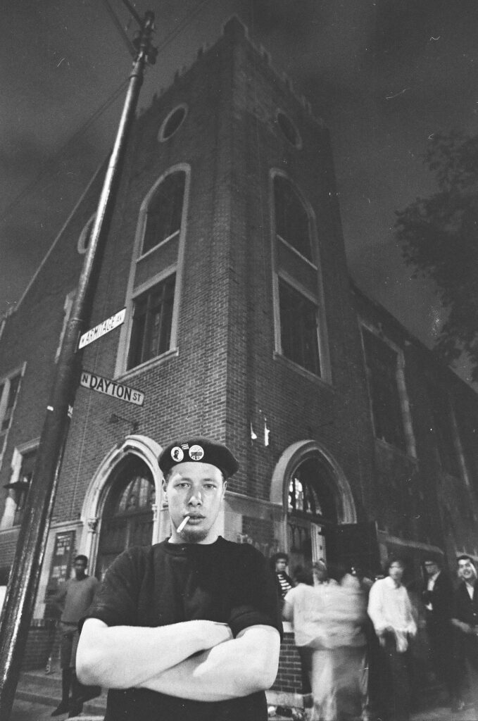 Black and white picture of Jose Cha Cha Jimenez standing outside of the Armitage Avenue Methodist Church wearing a black shirt and a black barrette with his arms crossed and cigarettes in his mouth.