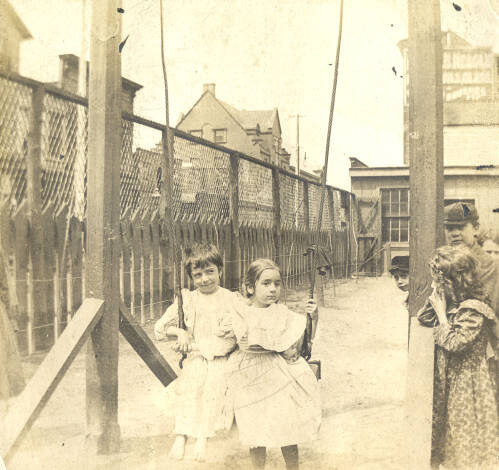 Two-girls-on-the-Hull-House-playground A sepia-colored photograph of two little girls sharing a swing. The swing is a plank of wood tied with rope to two very thick and tall wooden posts. There are four more children lined up, leaning on the left pole, presumably waiting for their turn on the swing. In the background there are two fences, one is made of wooden planks with pointed tops, the other is a chain-link fence about double the height. The two girls on the swing are barefoot and in long dresses. One has short hair and has a grin on her face, the other girl has long hair that is pulled back.and a blank expression.