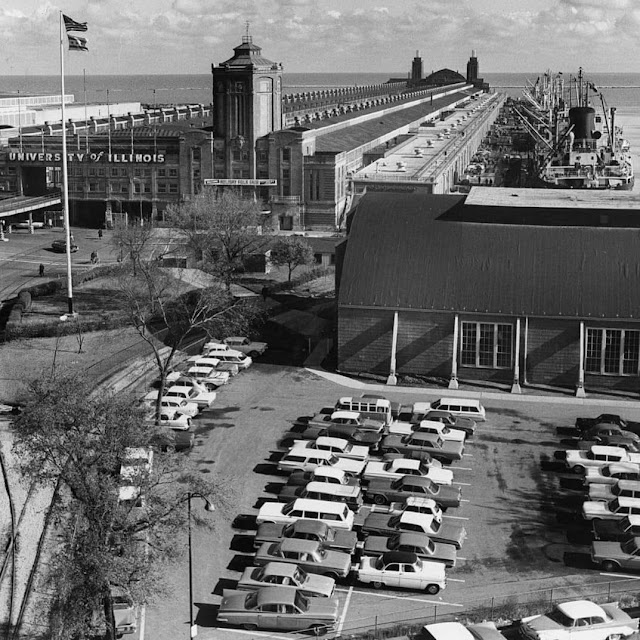 navy pier photo 1 a black and white photo of the Navy Pier Chicago in 1961. There is a long, low building that extends into Lake Michigan with the words UNIVERSITY OF ILLINOIS on the front. There is also a courtyard with a tall flagpole flying the American and Chicago flags. In the foreground there is a large parking lot full of convertables