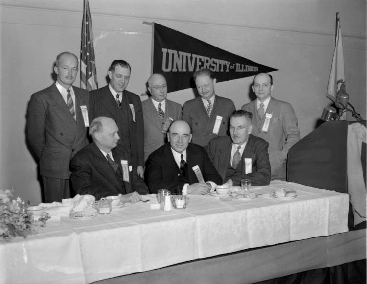 Citizens' Committee A black and white photo with three older men sitting at a long, cloth-covered table. Behind them are five men standing with their arms behind their backs. Most of the men are looking into the camera, but some men are looking at the man sitting in the middle. They look to be in their fifties or older. Behind them is an American flag and a triangle banner with "University of Illinois" written on it. On the far right of the photograph is a wooden podium with a microphone set up.